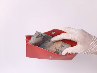 Beyaz lateks eldivenli bir elin içinde kırmızı bir çanta, içinde para, 2000 IDR ve Endonezya banknotları var. Beyaz arka planda izole edilmiş. Yan görünüm.