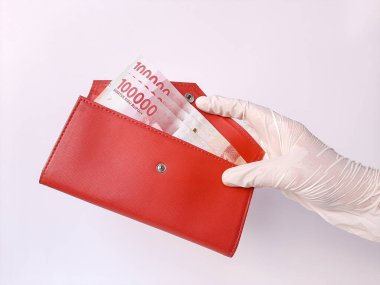 Beyaz lateks eldivenli bir el, içinde 100 bin IDR, Endonezya banknotları olan kırmızı bir çanta taşıyor. Beyaz arka planda izole edilmiş. Yan görünüm.