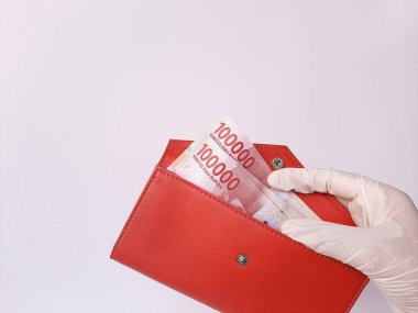 Beyaz lateks eldivenli bir el, içinde 100 bin IDR, Endonezya banknotları olan kırmızı bir çanta taşıyor. Beyaz arka planda izole edilmiş. Yan görünüm.