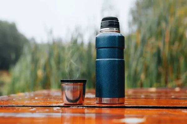 Thermos and mug with hot drink standing on wet wooden table after rain outdoors, close-up. Steam rises from thermo mug. Warming drink from hiking flask in cold, rainy weather.