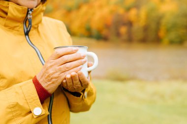 Yaşlı bir kadının elinde sıcak çay ya da kahveyle demir bir bardak tutarken, emeklinin dışarıda bir sonbahar parkında kendini ısıttığı yakın plan. Soğuk sonbahar havasında ılık içecek, boşluğu kopyala.