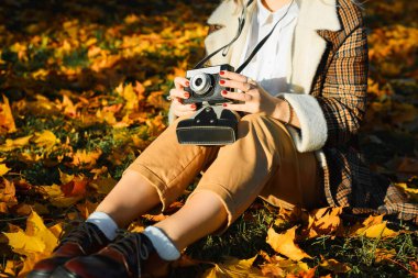 Sonbaharda dökülen yaprakların üzerinde otururken elinde eski moda film kamerası tutan bohem elbiseli genç bayan hippi fotoğrafçı, yakın plan. Hobi ve yaşam tarzı konsepti.