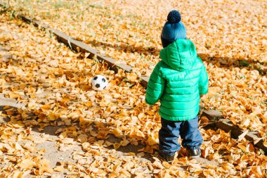 Kalın elbiseli bir çocuğun futbol topuyla sonbahar parkında futbol oynaması. Seçici olarak erkek bebeğe odaklan. Aktif spor, çocukluk.