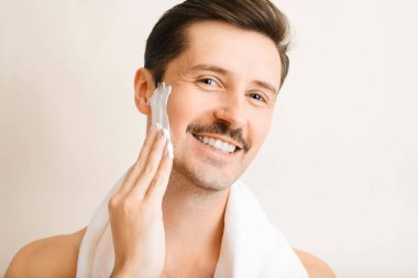 Yakışıklı, tıraşsız genç adam nemlendirici sürüyor, yüzüne köpük sürüyor ve kameraya bakıyor. Omuzlarında havlu olan mutlu bir adamın portresi günlük kişisel bakım ve kapalı alanda güzellik gösterisi yapıyor..