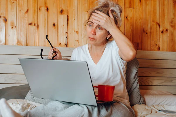 Sick adult woman with headache and virus working on laptop while sitting on bed with mug of tea in living room indoors. Freelancer woman tired with flu alone at home.