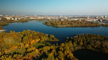 Sonbahar parkında uçuş. Sarı sonbahar yapraklı ağaçlar görülebilir. Ufukta mavi bir gökyüzü ve şehir evleri var. Park gölü görülebilir. Hava fotoğrafçılığı.