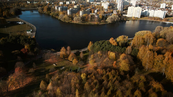 Полет над осенним парком. Парк на берегу большого озера. Городские дома. Видны деревья с жёлтыми осенними листьями. Аэрофотосъемка.