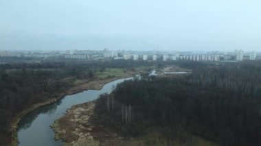Şehir parkının içindeki nehir. Sonbaharın sonu, bulutlu. Yapraksız ağaçlar ve nehir kıyısında kurumuş sazlıklar. Şehir ufukta görünüyor. Hava fotoğrafçılığı.