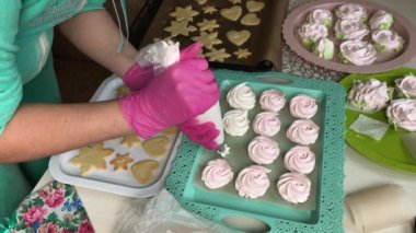 Kadın marşmelovlu sandviç hazırlıyor. Kurabiyelerin üzerine şekerleme koyuyor. Yakınlarda kalp şeklinde kurabiyeler ve kar taneleri var..