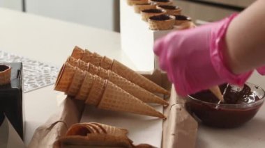 Bir kadın waffle külahlarını sıvı çikolatayla kaplıyor ve tezgahın üzerine koyuyor. Çikolata ve lokumla boynuz yapıyor. Yakın plan..