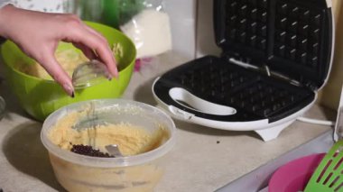 Bir kadın waffle hamuruna çikolata damlası ekler. Malzemeleri karıştırıyor. Yanında bir waffle ızgarası var. Yakın plan..