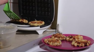 Bir kadın ısıtılmış waffle ızgarasını açar ve bitmiş waffleları çıkarır. Tabağa koyuyor. Kahvaltı hazırlığı. Yakın plan.