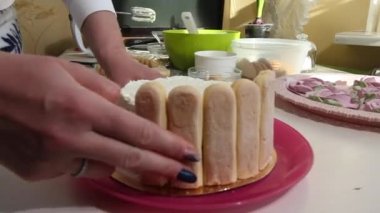 Bir kadın pastayı Savoiardi kurabiyeleriyle kaplıyor. Pastanın malzemeleri masanın üzerine serildi. Savoiardi kurabiyeli kek ve marshmallow gülleri..