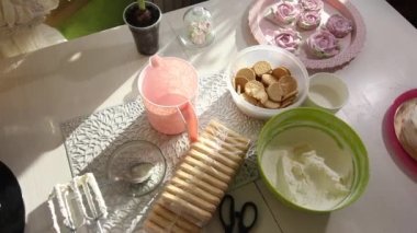 Bir kadın pasta hazırlıyor. Malzemeler masanın üstünde. İçinde Savoiardi kurabiyeleri ve marshmallow gülleri olan kek..