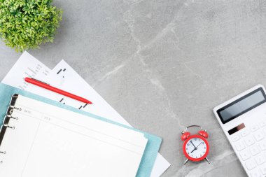 Metal bir kalem, hesap makinesi, çalar saat, borsa çizelgeleri ve gri mermer arka planda saksıda bir çiçekle kaplı, yumuşak mavi kapaklı bir defter. Ofis masası konsepti. Üst görünüm.