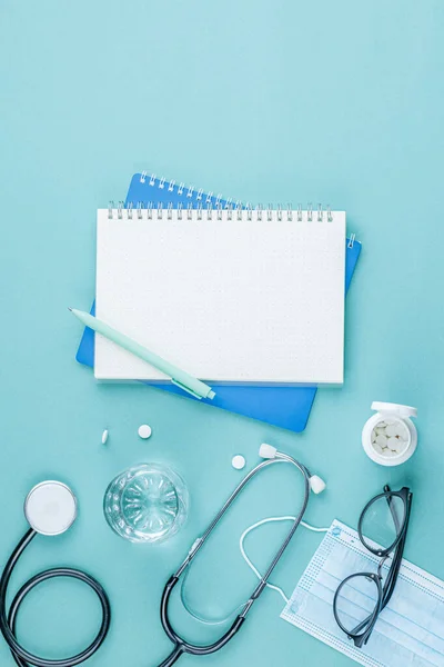 Drugs and glass of water on green medical background top view. Stethoscope and notebook with a pen at the workplace of a doctor. Nurse desktop concept. Vertical photo.