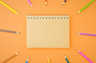 Merkezde, turuncu arkaplanda daire şeklinde dizilmiş renkli kalemlerden yapılmış spiral el işi defter var. Üst görünüm.