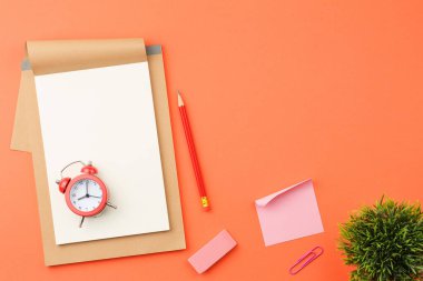 Beyaz sayfalı bir defter, kırmızı alarm saati ve kalem, lastik bant, ataç ve turuncu izole arkaplan görünümlü not kağıdı.
