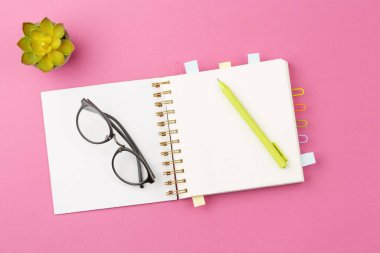 Open spiral notebook with bookmarks from paper clips and leaves for notes, pen, glasses and a flower in a pot on a pink background. Top view.
