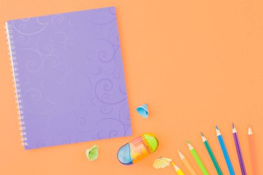 Spiral purple notbook with colored pencils sharpener and shavings on an orange background. Top view, copy space.