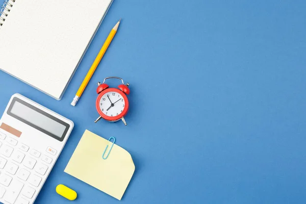 Spiral notebook with pencil, calculator, note sheet, paper clip, red alarm clock and rubber band on blue isolated background. Office concept. Top view.