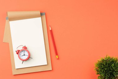 Beyaz çarşaflı el işi defter, kırmızı alarm saati, yeşil yapraklar, turuncu izole arka plan görünümlü bitki ve kalem..