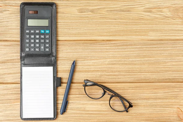 Calculator with a notebook in a leather case with a dark blue pen and black glasses on a light wooden background flat lay.