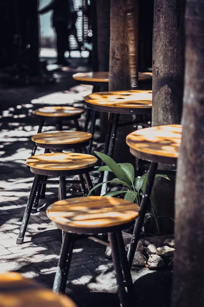 Vertical atmospheric warm summer cover photo, wooden stools chairs in outdoor cafe on veranda in garden, bright rays sun beam through trees. Positive good calm mood concept, for wallpaper screensaver