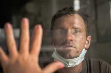 Beyaz koruyucu maske takan, camdan dışarı bakan, gözlerindeki stresten korkan, evdeki hastalıktan kurtulan, COVID 19 Coronavirus salgınından dolayı kendini izole eden bir adam.