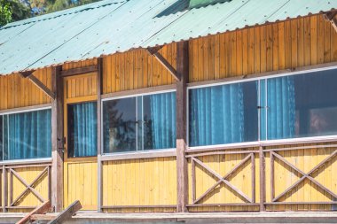 Dış görünüş. Tahta sarısı kahverengi tahta kumaş, kır evi, panoramik pencere camı yansıması, perdeler içeri. Yaz günü sıcak hava. Konforlu ev içleri, boş zaman, ekoloji binası