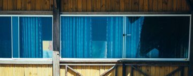 Dış görünüş. Tahta sarısı kahverengi tahta kumaş, kır evi, panoramik pencere camı yansıması, perdeler içeri. Yaz günü sıcak hava. Konforlu ev içleri, boş zaman, ekoloji binası