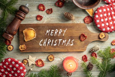 Kozalaklı dallar, şenlik ve mutfak aksesuarları ve ahşap zemin üzerinde Mutlu Noeller yazan kahverengi bir kesme tahtası. Üst görünüm.