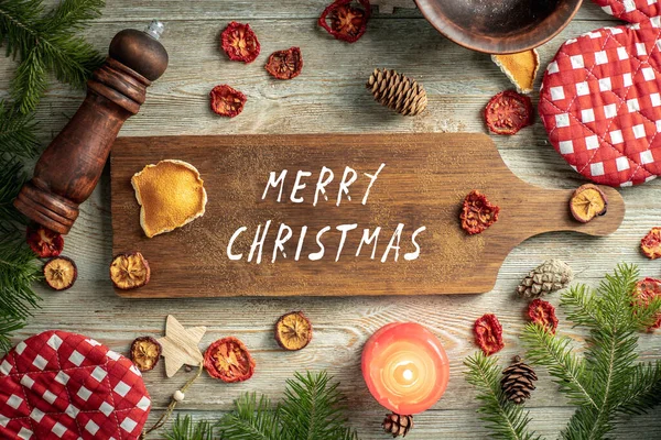 Kozalaklı dallar, şenlik ve mutfak aksesuarları ve ahşap zemin üzerinde Mutlu Noeller yazan kahverengi bir kesme tahtası. Üst görünüm.
