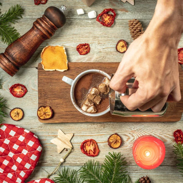 手は白いカップにホットチョコレートを注いでいます 木製のテーブルの周りにお祝いのアクセサリーです 居心地の良いクリスマスと新年の気分の概念 — ストック写真