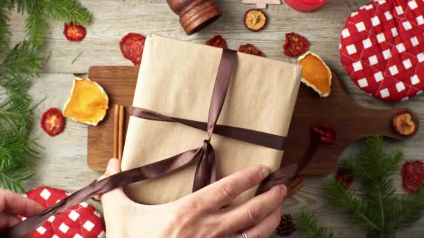 Las manos están atando una cinta marrón en un regalo envuelto en papel artesanal. Concepto de preparación para el Año Nuevo y Navidad, embalaje de regalos para las vacaciones. Vista superior — Vídeos de Stock