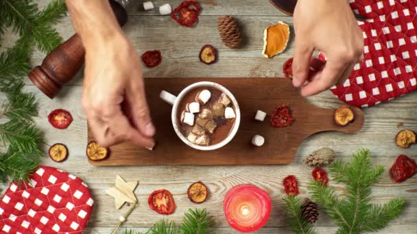 Hände bestreuen heiße Schokolade mit Marshmallows und rühren sie mit einer Zimtstange um. Auf einem Holztisch liegen festliche Accessoires herum. Konzept der Weihnachts- und Neujahrsstimmung — Stockvideo