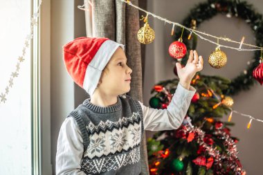 Kırmızı Noel Baba şapkalı şirin çocuk Noel ağacının yanındaki pencerede oturuyor ve evdeki şenlikli süslemelere bakıyor. Yeni yıl atmosferi ve tatil havası kavramı.