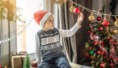 Kırmızı Noel Baba şapkalı şirin çocuk Noel ağacının yanındaki pencerede oturuyor ve evdeki şenlikli süslemelere bakıyor. Yeni yıl atmosferi ve tatil havası kavramı.
