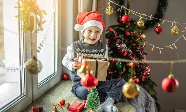赤いサンタの帽子の幸せな少年は 休日のために装飾された部屋のクリスマスツリーの隣の窓に座っていて 彼の手にギフトボックスを持っています 新年の雰囲気とお祝いムードの概念 — ストック写真