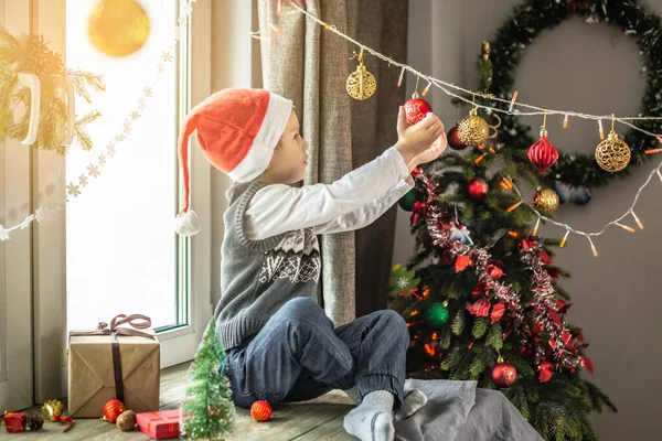 赤いサンタの帽子のかわいい男の子はクリスマスツリーの隣のウィンドウに座っており 自宅でお祝いの装飾を見ています 新年の雰囲気と休日の気分の概念 — ストック写真