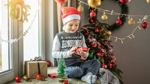 赤いサンタの帽子のかわいい男の子は 装飾された部屋の窓の隣に座っていて 彼の手に小さなオープンギフトボックスを持っています お正月とクリスマス気分のコンセプト — ストック写真