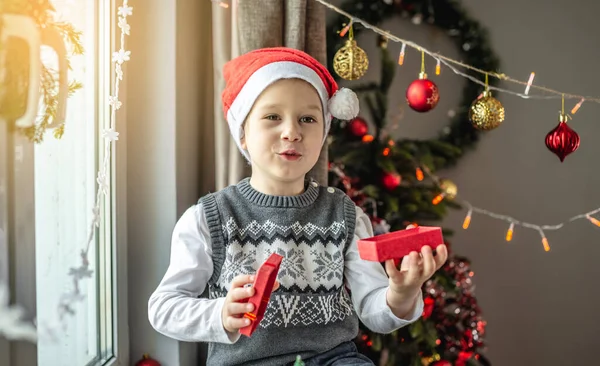 赤いサンタの帽子のかわいい男の子は装飾された部屋の窓のそばに座っていて 彼の手に小さなオープンギフトボックスを持っています お正月とクリスマス気分のコンセプト — ストック写真