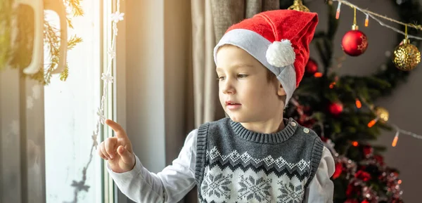 赤いサンタの帽子のかわいい男の子はクリスマスツリーの隣のウィンドウに座っており 自宅でお祝いの装飾を見ています 新年の雰囲気と休日の気分の概念 — ストック写真