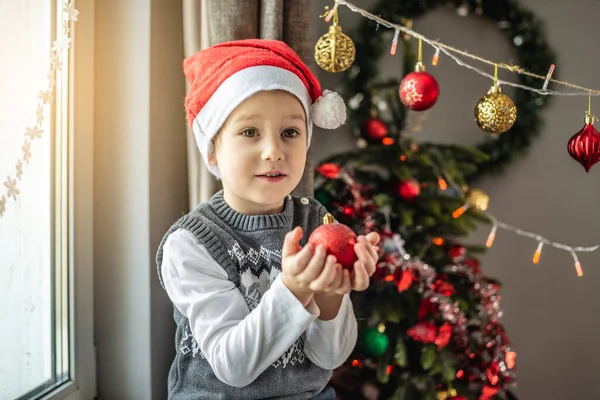 赤いサンタの帽子のかわいい男の子は装飾された部屋のクリスマスツリーの隣に座っていて 彼の手にクリスマスボールを持っています お正月気分のコンセプト — ストック写真