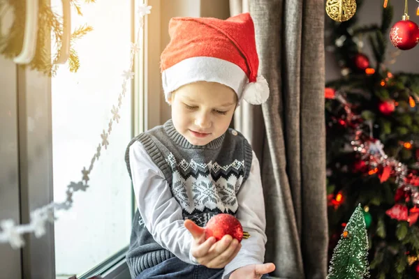 サンタの帽子のかわいい男の子は装飾された部屋のクリスマスツリーの隣に座っていて 彼の手にクリスマスのおもちゃを持っています 正月の準備の概念は 休日の雰囲気 — ストック写真