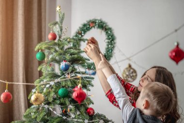 Anne ve çocuğu Noel ağacını oyuncaklar ve toplarla süslüyorlar. Yeni yıla aile hazırlığı, rahat atmosfer ve şenlik havası kavramı.