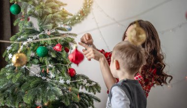 Anne ve çocuğu Noel ağacını oyuncaklar ve toplarla süslüyorlar. Yeni yıla aile hazırlığı, rahat atmosfer ve şenlik havası kavramı.