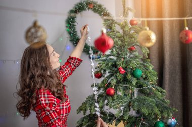 Kırmızı elbiseli genç kadın Noel ağacını oyuncaklar ve toplarla süslüyor. Yeni yıla hazırlık kavramı, rahat atmosfer ve şenlik havası.