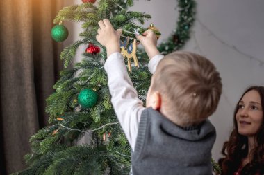 Anne ve çocuğu Noel ağacını oyuncaklar ve toplarla süslüyorlar. Yeni yıla aile hazırlığı, rahat atmosfer ve şenlik havası kavramı.