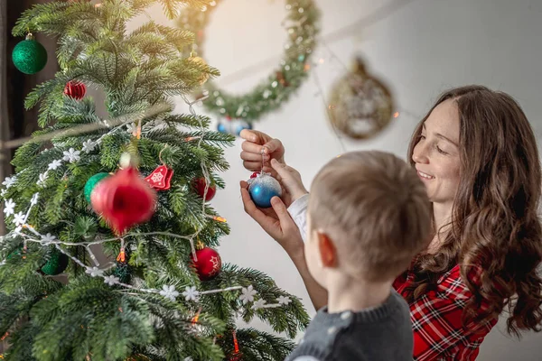 Anne ve çocuğu Noel ağacını oyuncaklar ve toplarla süslüyorlar. Yeni yıla aile hazırlığı, rahat atmosfer ve şenlik havası kavramı.
